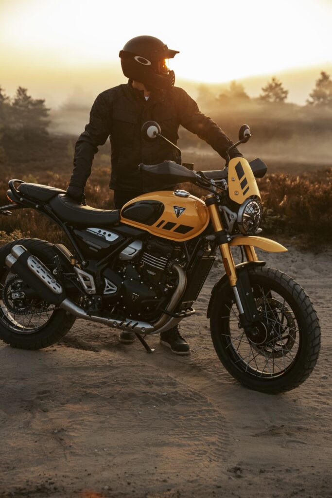 Triumph Unveils the All New 2026 Scrambler 400 XC 6 a man weating a helmet stands behind a motorcycle on a dirt road