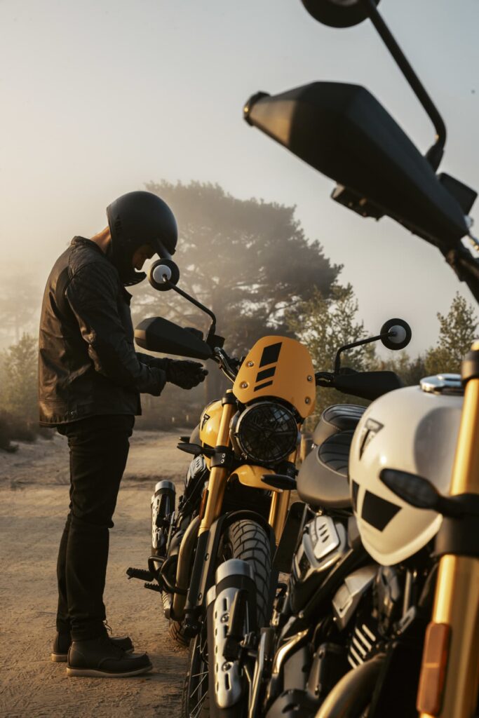 Triumph Unveils the All New 2026 Scrambler 400 XC 4 a man stands alongside a motorcycle on a dirt road